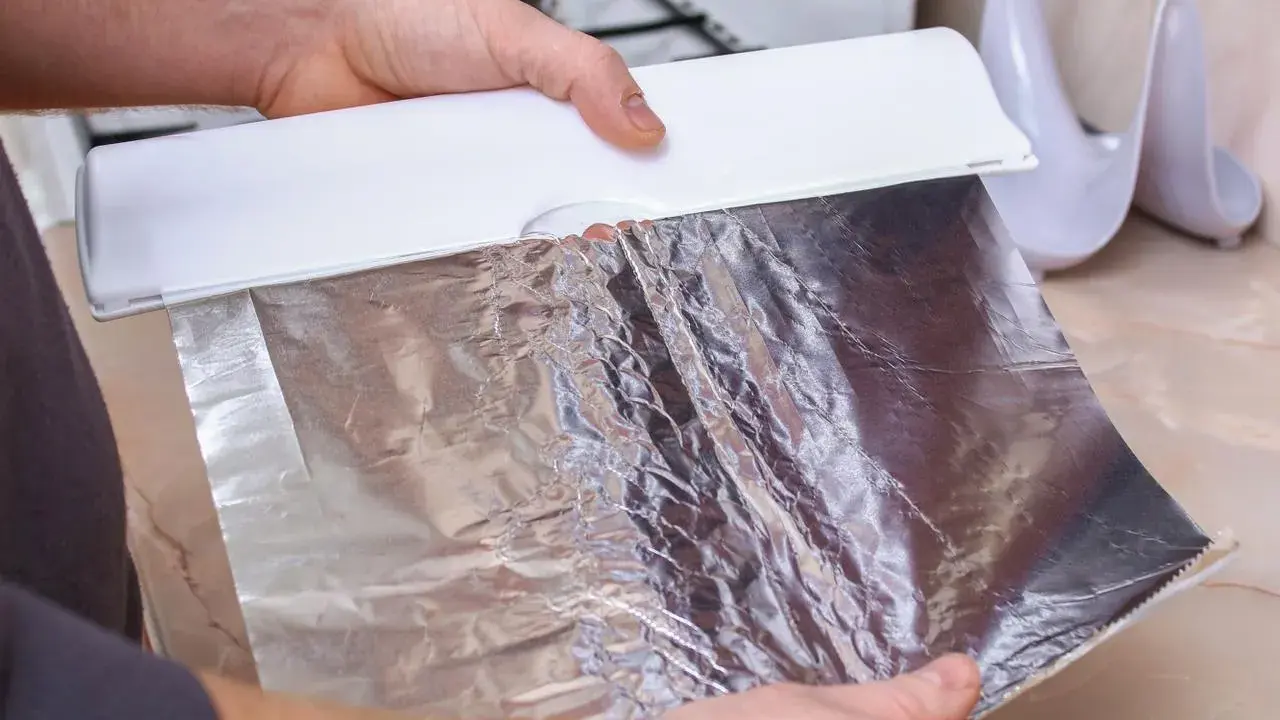 Ręce trzymają biały dozownik z folią aluminiową. Zastanawiasz się, która folia aluminiowa strona do pieczenia? Ta błyszcząca strona odbija ciepło.
