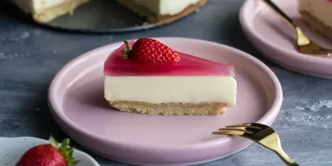 Pyszny placek z galaretką i truskawką na różowym talerzyku. Obok całe ciasto i kolejna porcja.