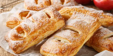 Chrupiące ciasto francuskie z jabłkami, posypane cukrem pudrem, czeka na spróbowanie. Obok leżą rumiane jabłka.