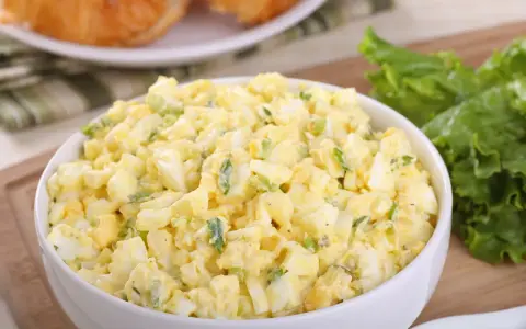 Pyszna sałatka z jajkiem, idealna na śniadanie lub lunch. Podana w białej misce, obok świeżych liści sałaty i chrupiących croissantów.
