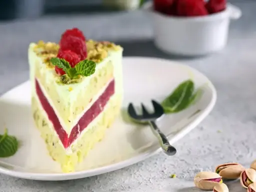 Pyszny tort pistacjowo malinowy z malinami i miętą na talerzu. Obok widoczne łuskane pistacje.