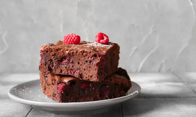 Dwa pyszne brownie z malinami, posypane cukrem pudrem, ułożone na szarym talerzyku.