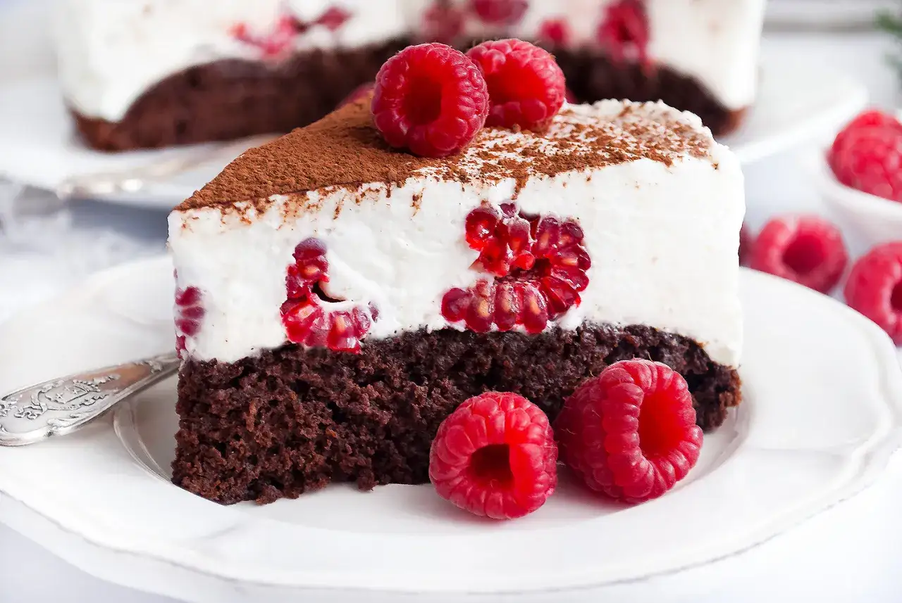 Pyszny brownie z malinami, udekorowany bitą śmietaną i kakao. Kilka świeżych malin zdobi wierzch ciasta.