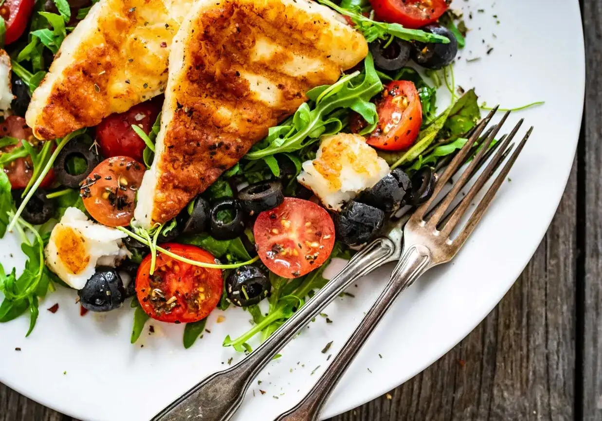 Pyszna sałatka z grillowanym halloumi, pomidorkami koktajlowymi i czarnymi oliwkami, podana na białym talerzu z widelcami.