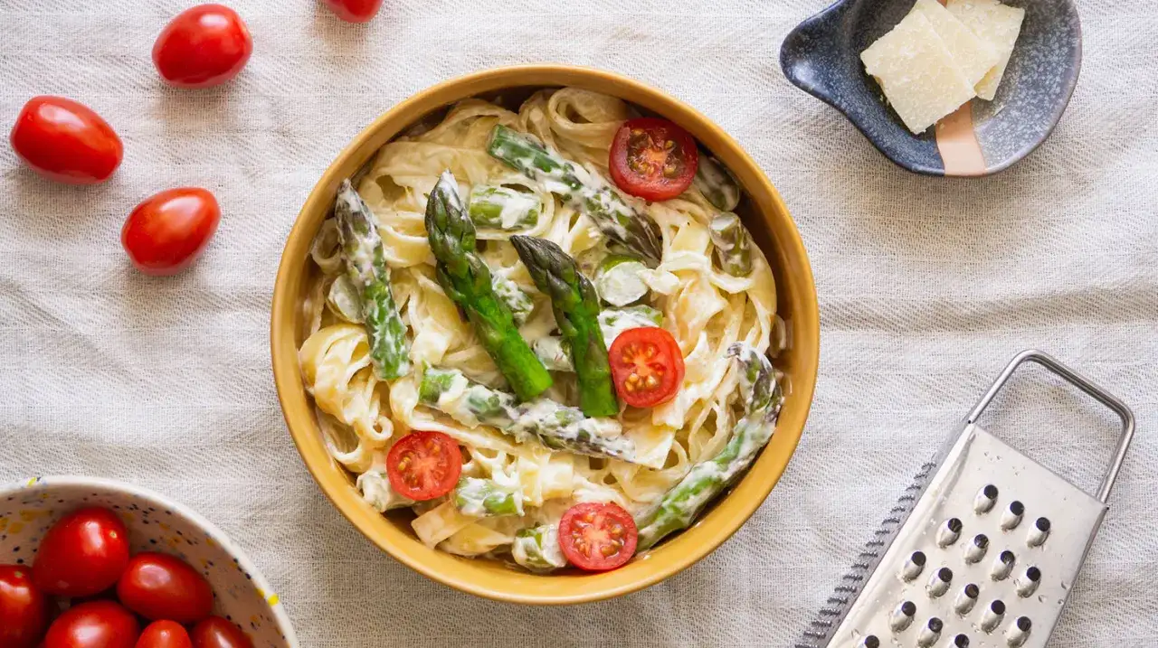 Pyszny makaron ze szparagami w kremowym sosie, udekorowany pomidorkami koktajlowymi. Obok miseczka z parmezanem i tarką.