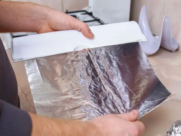 Ręce trzymają biały dozownik z folią aluminiową. Zastanawiasz się, która folia aluminiowa strona do pieczenia? Ta błyszcząca strona odbija ciepło.